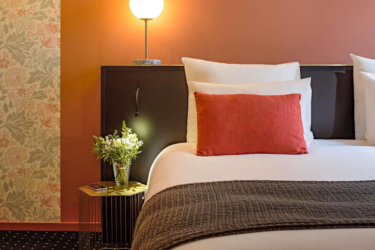 Una habitación de hotel con cama, almohadas, mesita de noche con lámpara y un florero con flores.