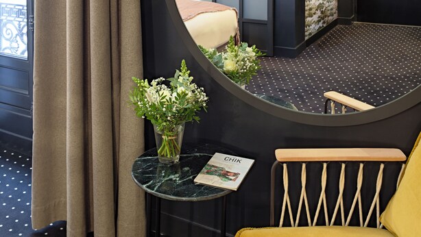 A room with a round mirror, a small table with a magazine, a yellow armchair, and a window with curtains.