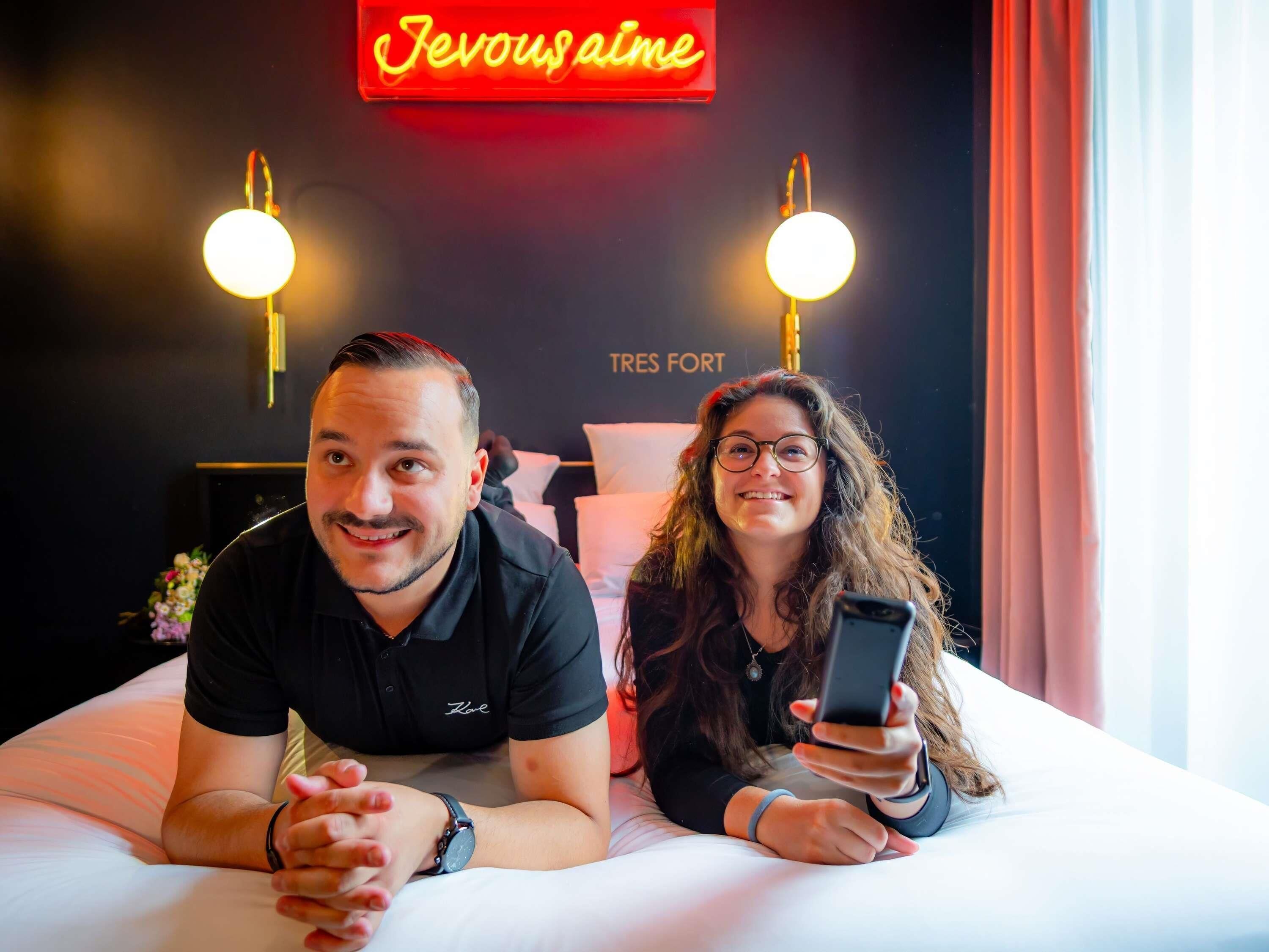 Two people lying on a bed in a hotel room with a neon sign above and two wall-mounted lamps.