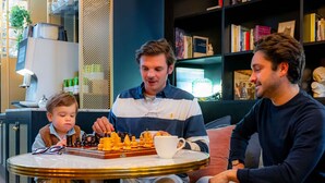 Een groep mensen zit rond een tafel met een schaakbord, in een kamer met een boekenkast en een lamp.