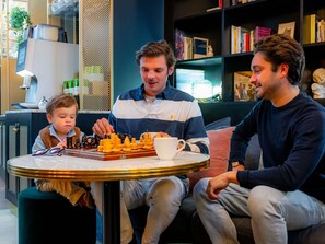 A group of people seated around a table with a chessboard, in a room with a bookshelf and a lamp.