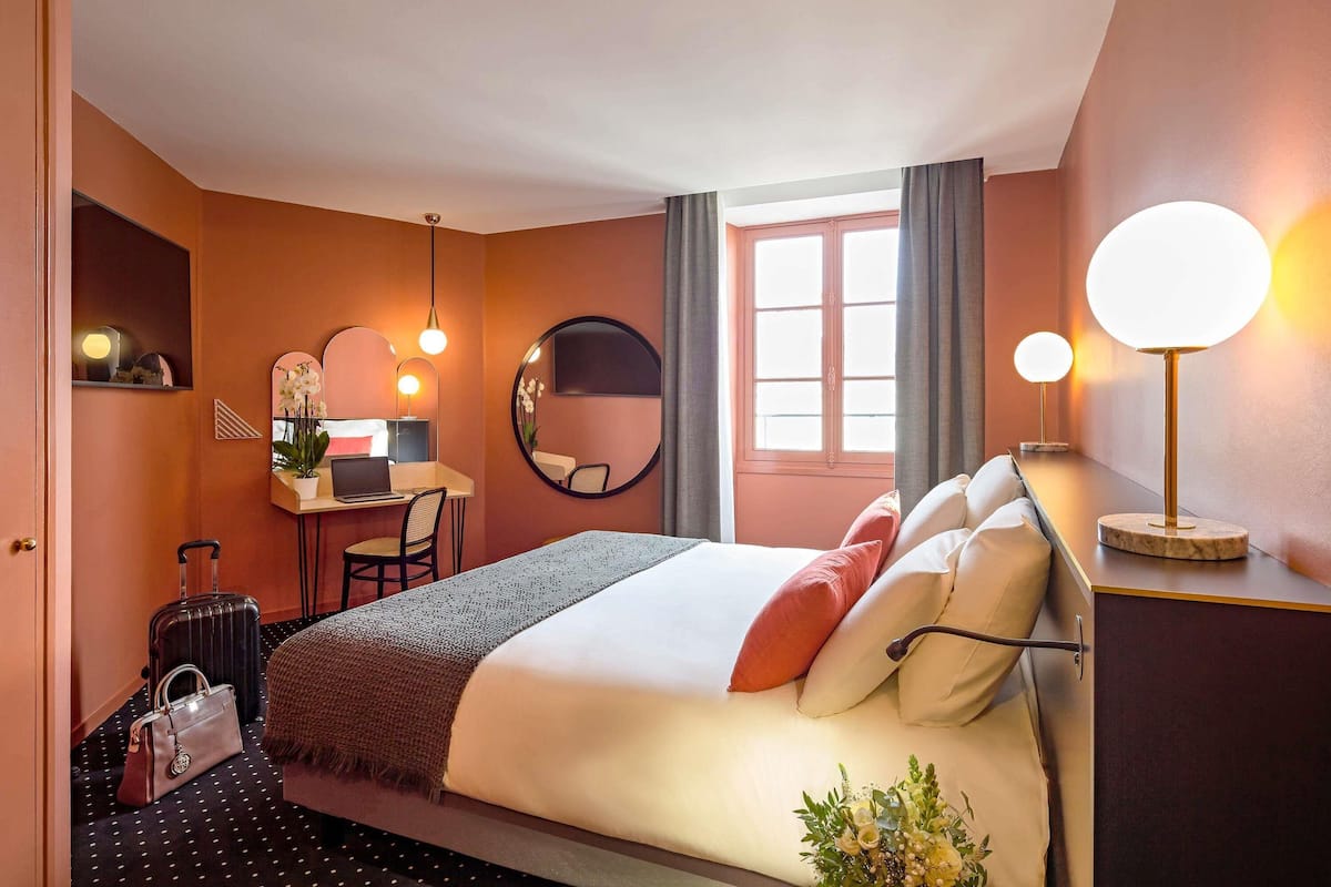 A hotel room with a bed, desk, chair, suitcase, and a lamp.