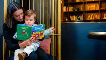 A person reading a book to a child in a room with a bookshelf and a decorative chair.