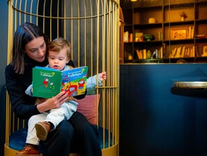 Een persoon leest een boek voor aan een kind in een kamer met een boekenkast en een decoratieve stoel.