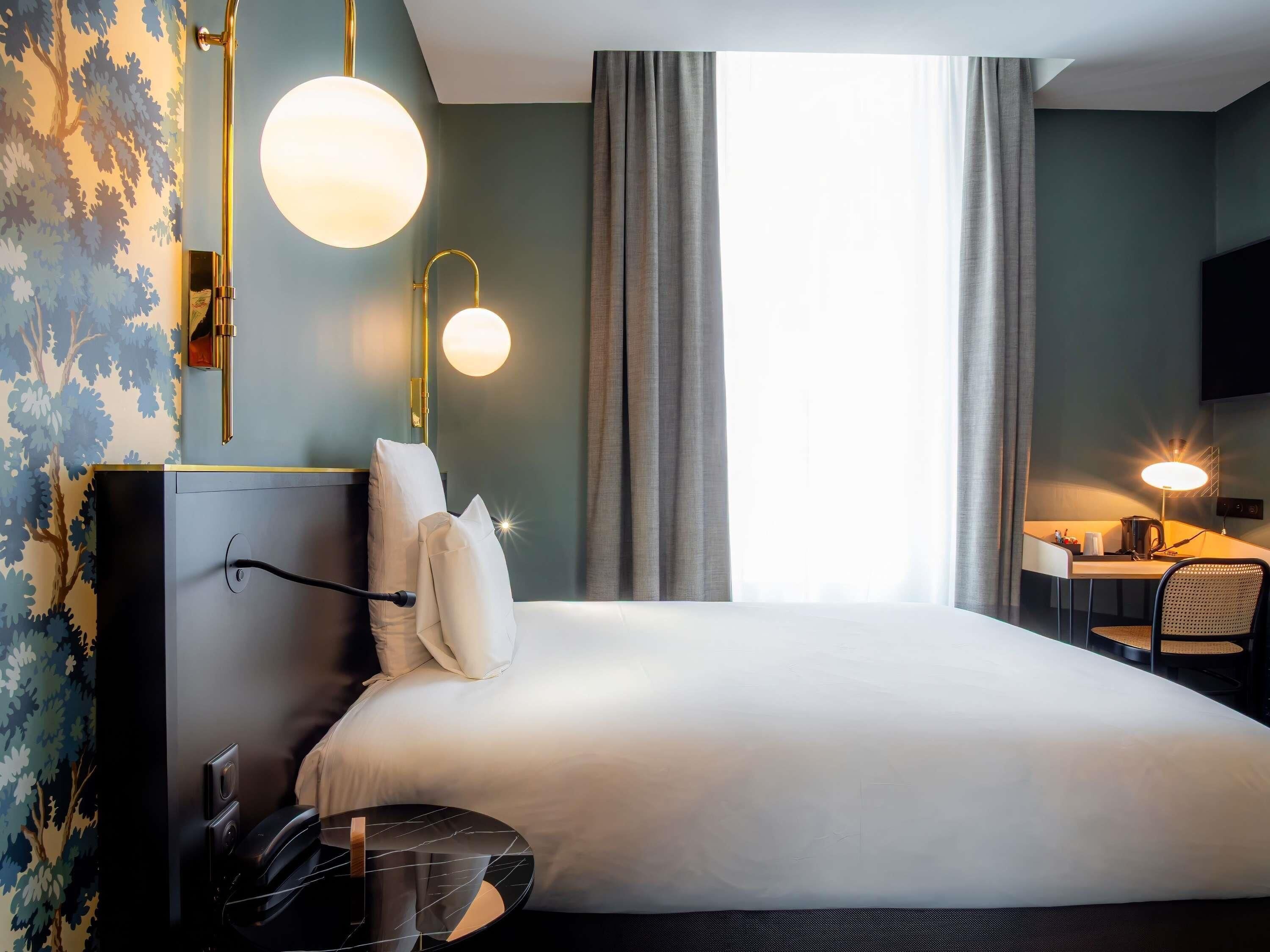 A hotel room with a bed, bedside table, lamp, and a view of the outdoors.
