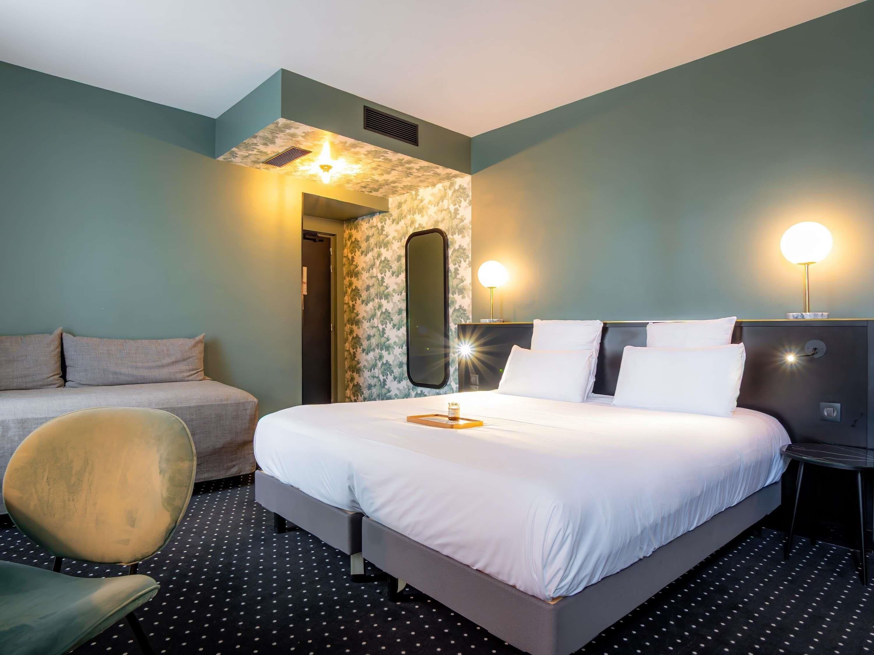 A hotel room with a bed, a sofa, a chair, and a nightstand with a lamp.