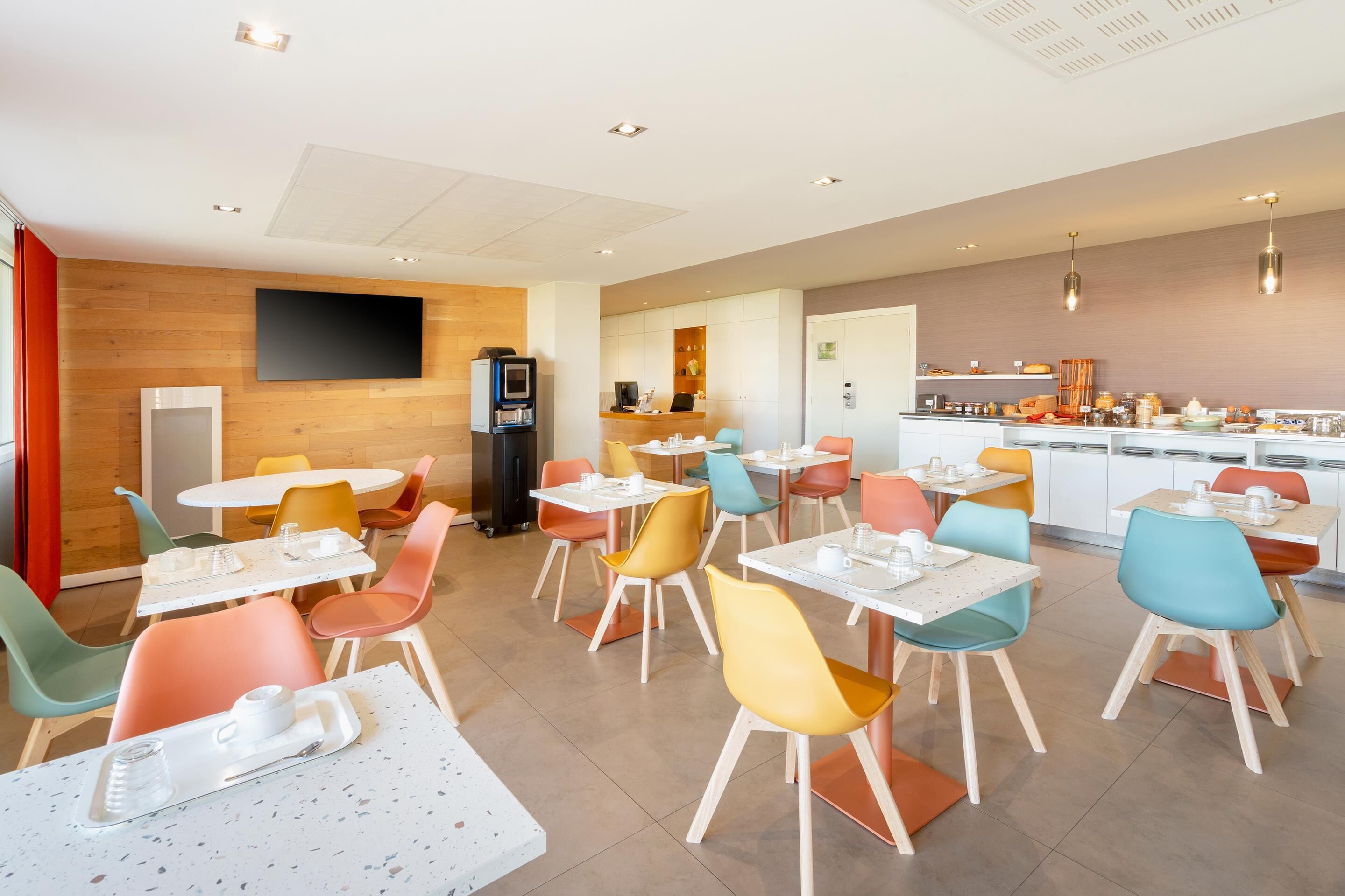 A modern dining area with colorful chairs and tables, a flat-screen TV, and a kitchen counter with various food items.
