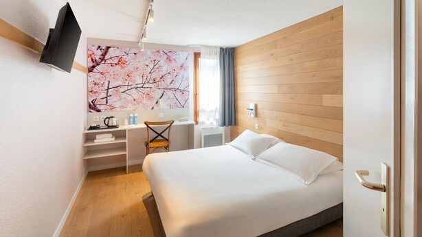 A hotel room with a bed, a desk with a chair, a TV mounted on the wall, and a window with curtains. - Sure Hotel by Best Western Rennes Chantepie (Chantepie)