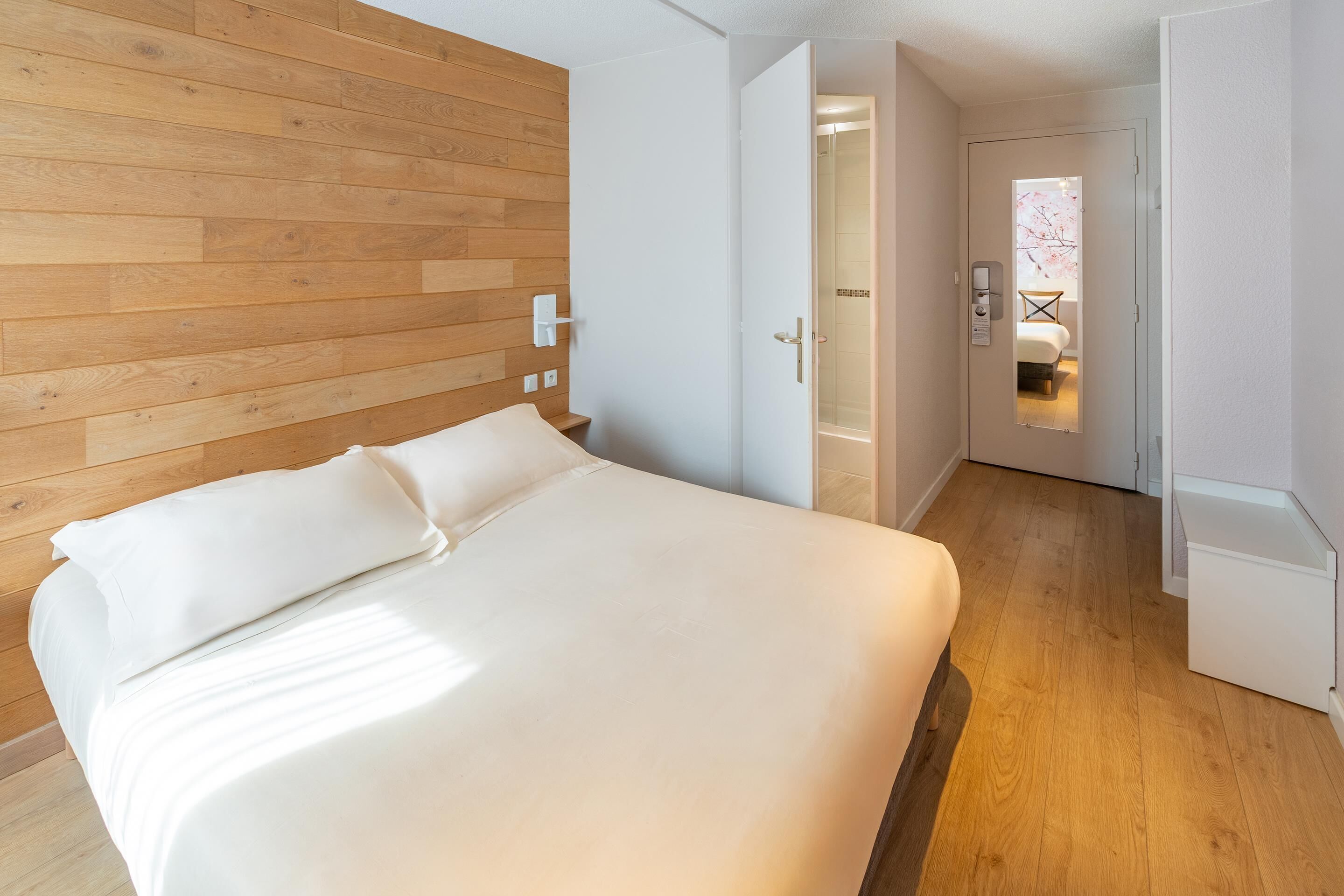 A modern hotel room with a wooden headboard, a large bed with white linens, a small bedside table, and a view into the bathroom through a glass door.
