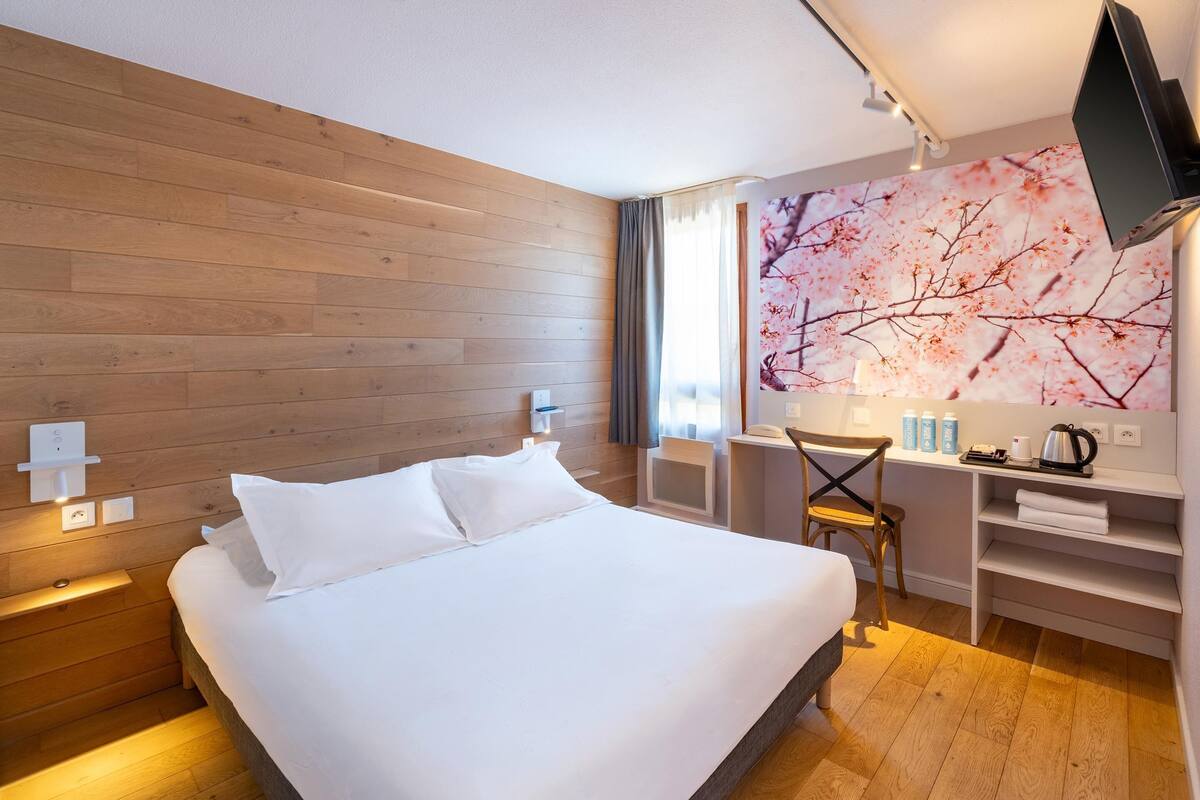 Ein Hotelzimmer mit einer Holzwand, einem Bett mit weißer Bettwäsche, einem Schreibtisch mit Stuhl, einem an der Wand montierten Fernseher und einem Fenster mit Vorhängen.