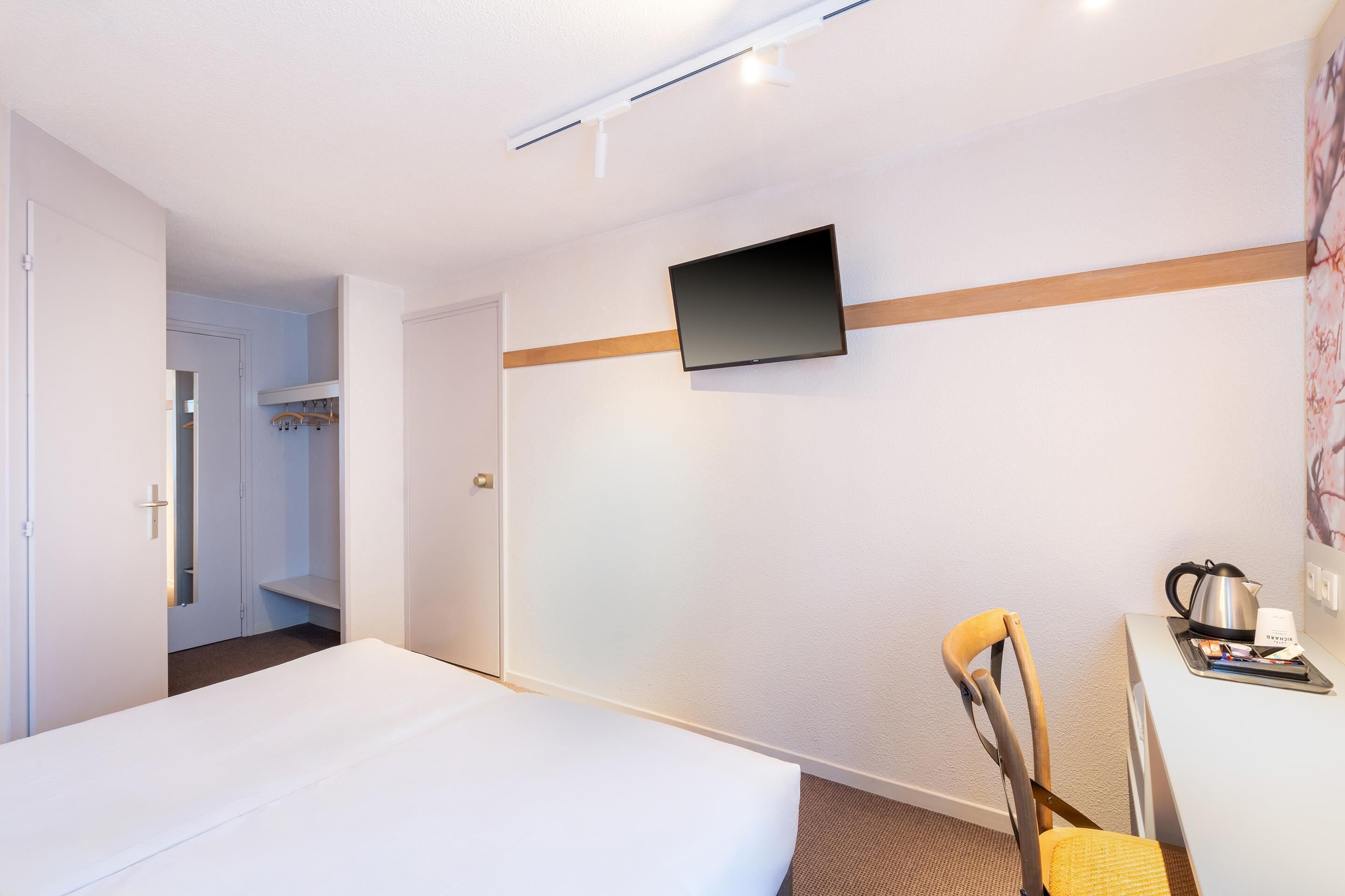 A hotel room with a bed, a desk with a kettle and a cup, a chair, a TV mounted on the wall, and a door leading to a bathroom.
