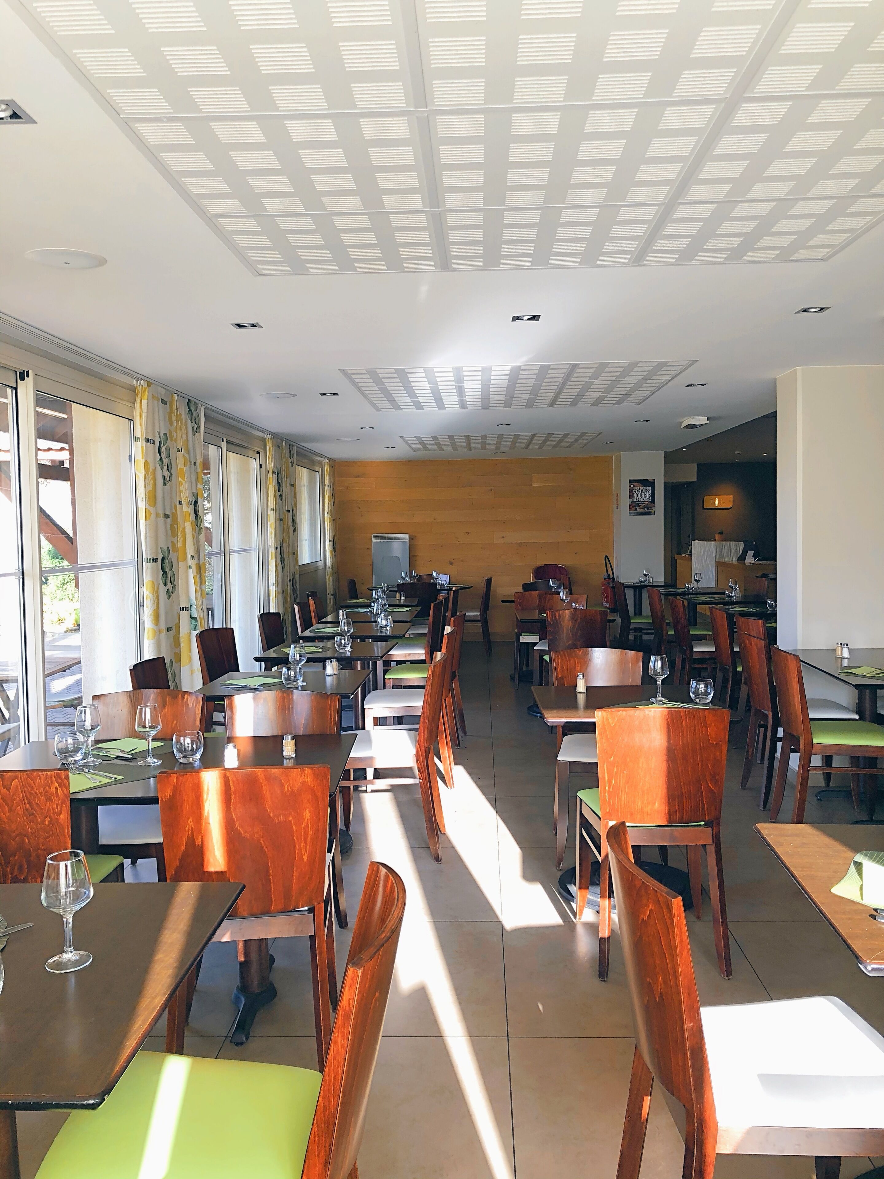 Een ruim restaurant met hoge plafonds, grote ramen en een modern interieur. Tafels staan netjes klaar met stoelen, en de wanden zijn bekleed met houten panelen.