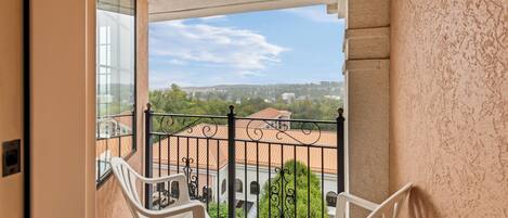 A balcony with two white plastic chairs, a wrought iron railing, and a view of buildings and greenery.
