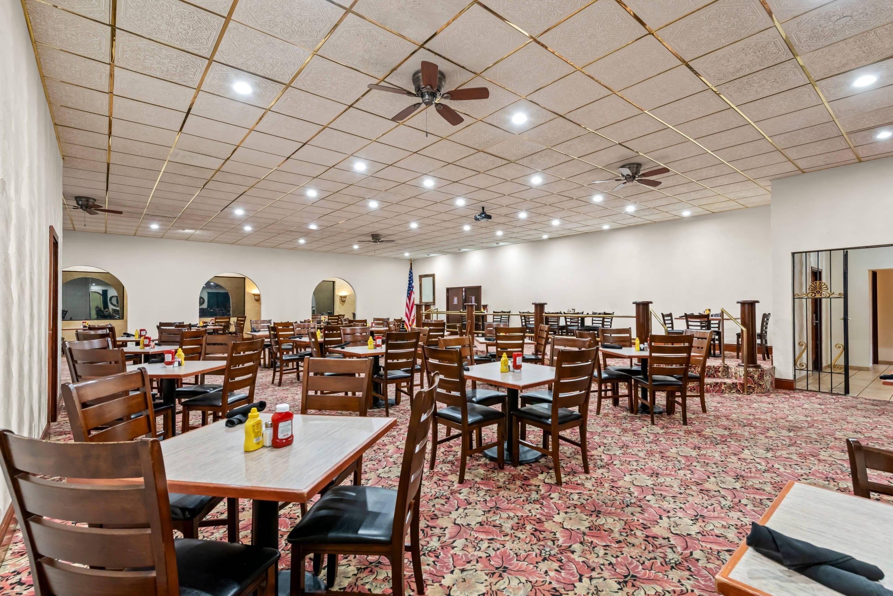 Un comedor amplio con varias mesas y sillas, ventiladores de techo y una alfombra estampada.