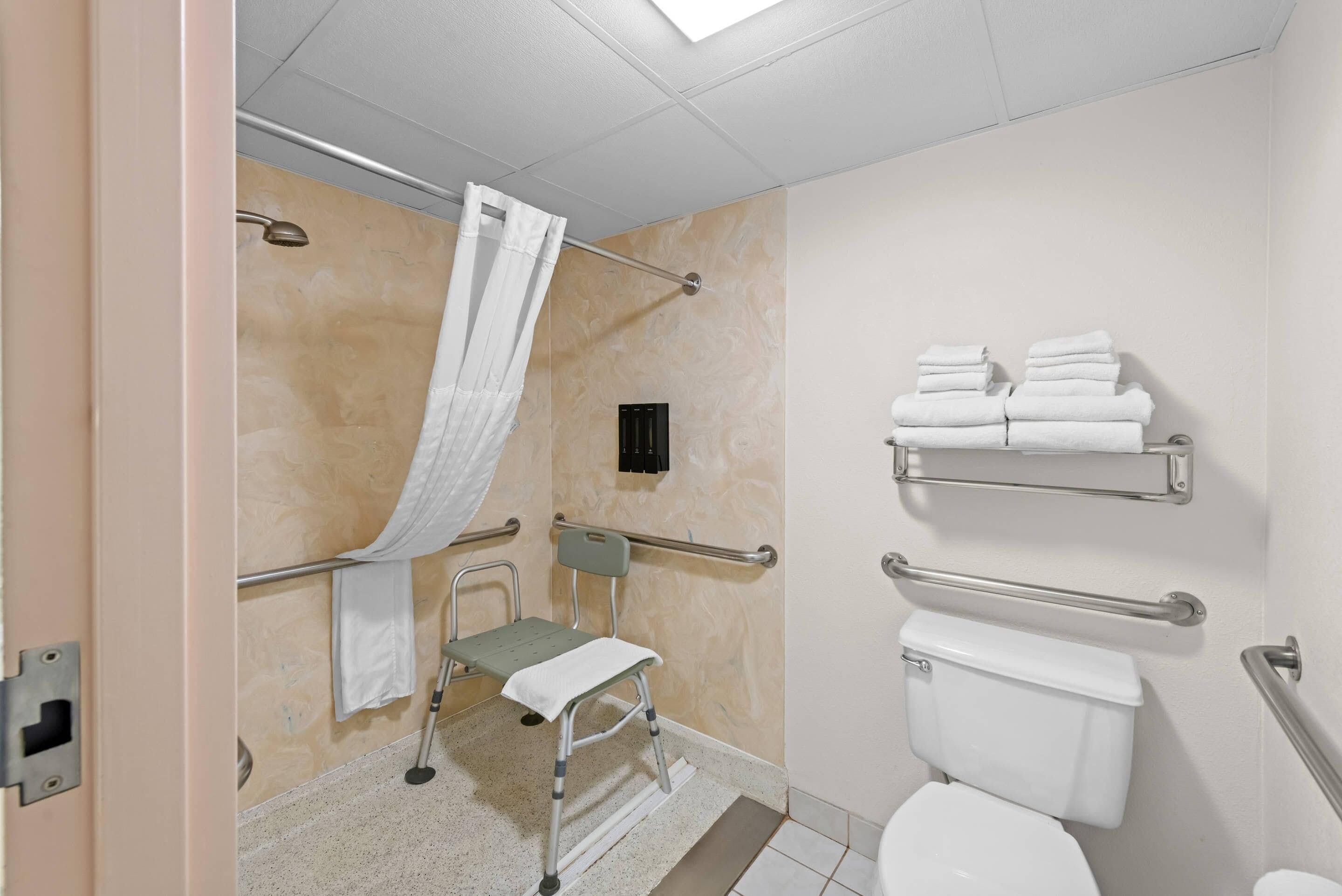 A bathroom with a shower, a toilet, a grab bar, a wheelchair, and a towel rack.