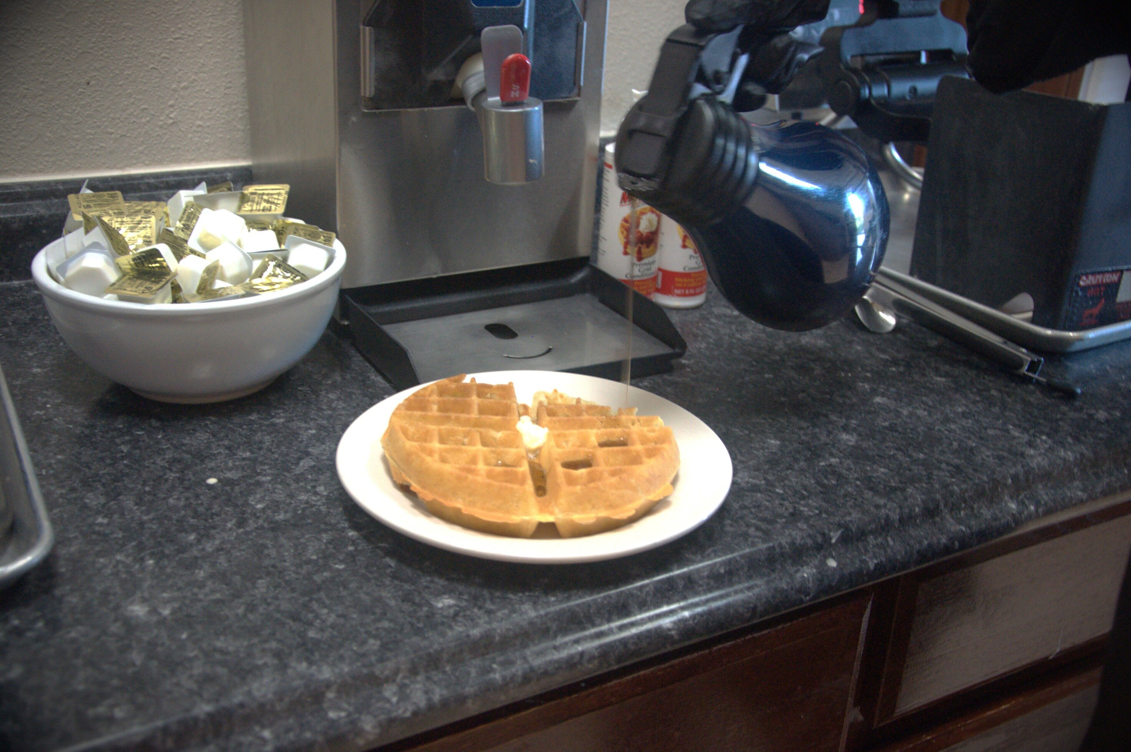 Un waffle en un plato, un tazón de monedas de oro y una cafetera sobre una encimera de cocina.