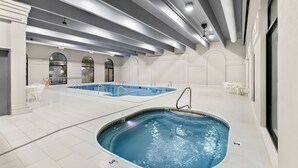 Una piscina interior con un pequeño jacuzzi, suelo de baldosas blancas y ventanas arqueadas.