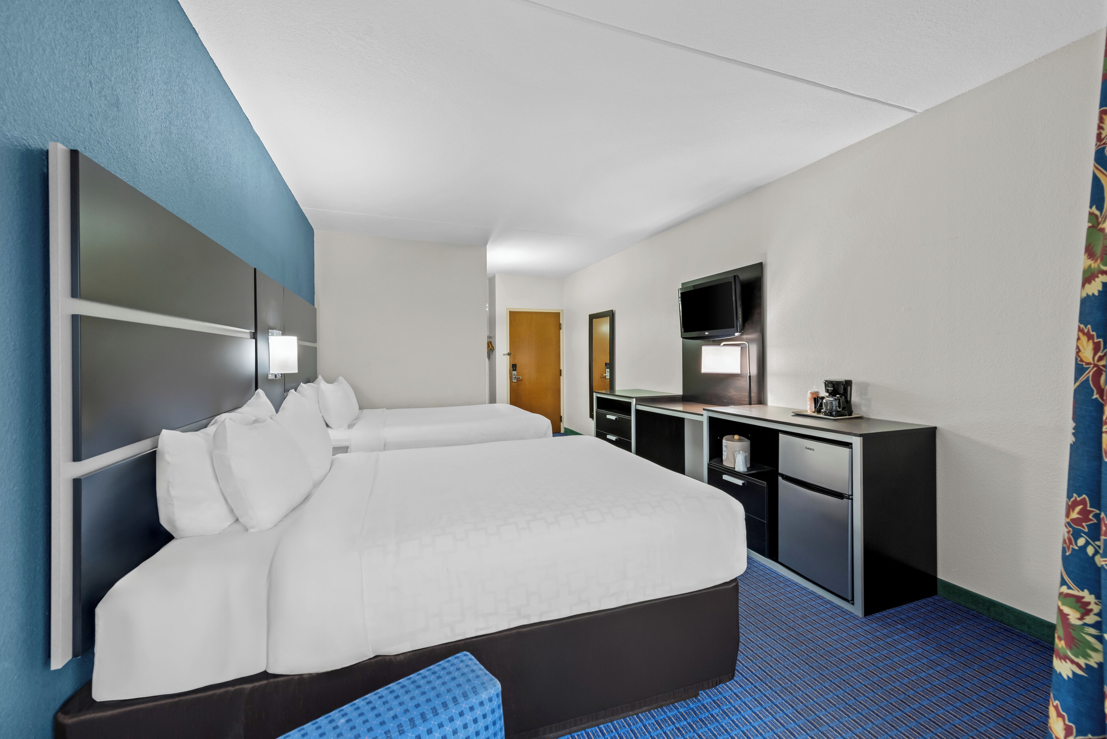 A hotel room with a large bed, a desk with a TV, and a minibar.