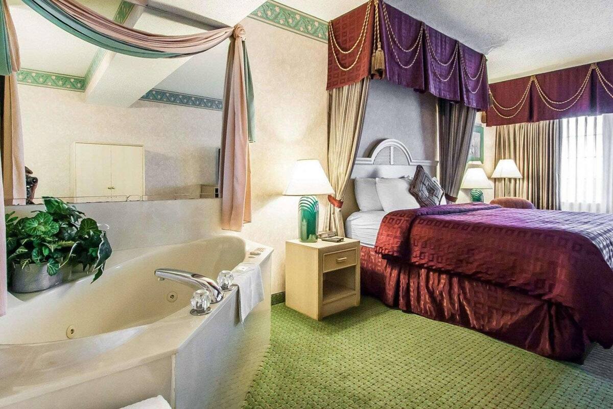Una habitación de hotel con una bañera grande, una cama con cubrecama rojo, dos lámparas de noche y una planta en maceta.