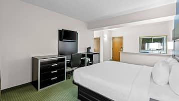 Quarto de hotel com cama, escrivaninha com gavetas, cadeira, televisão e espelho.