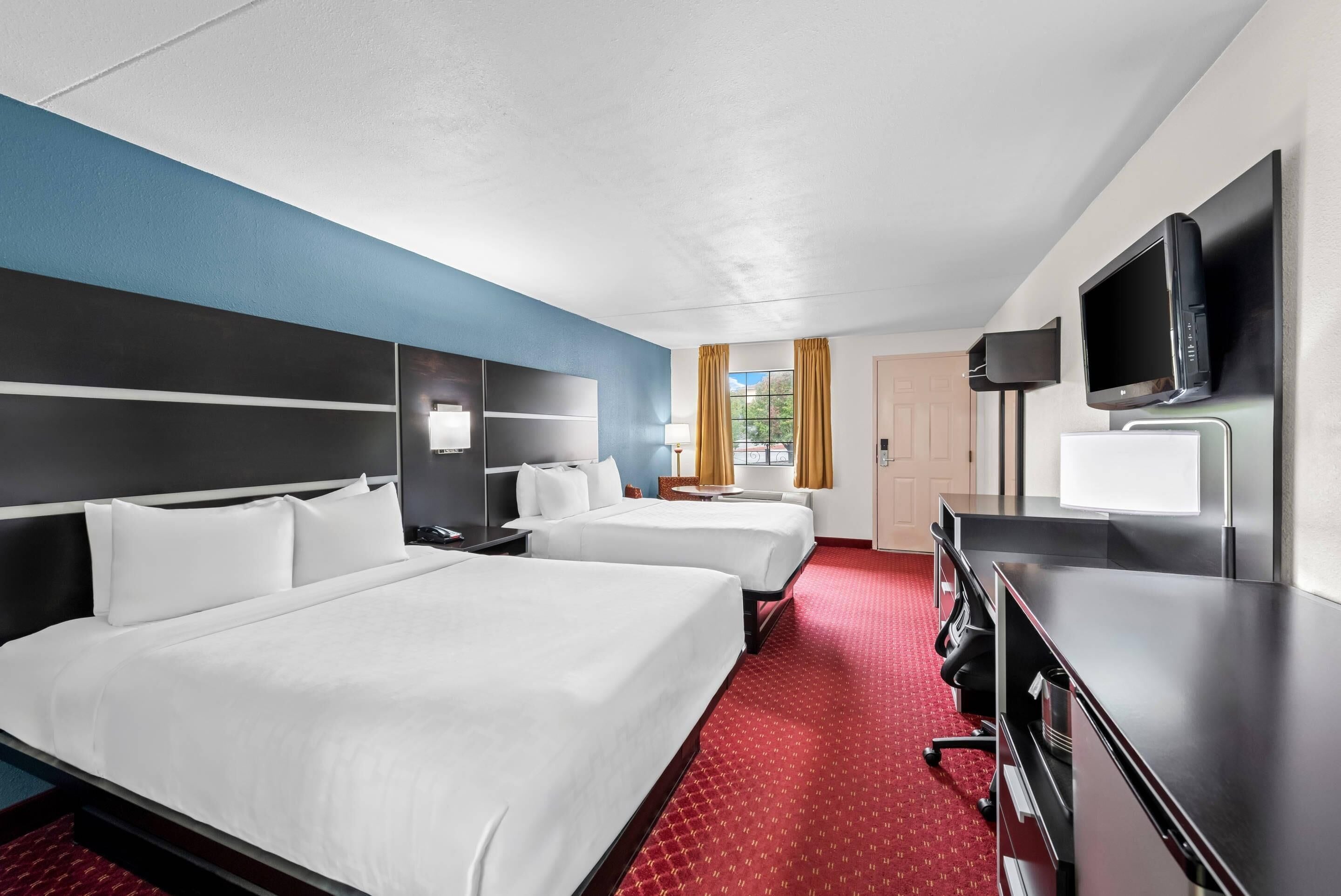 A hotel room with two beds, a desk, a TV, and a window with curtains.