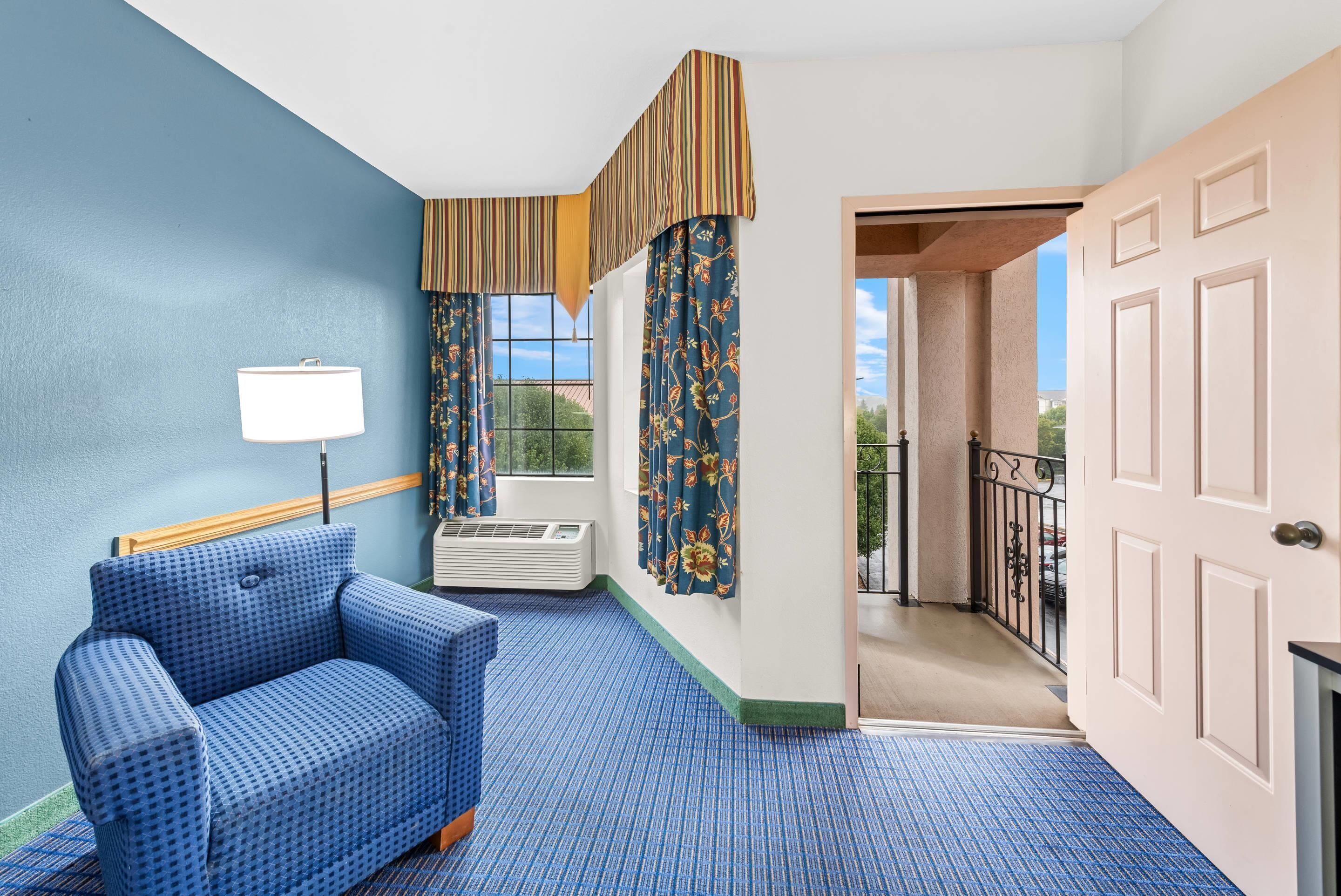 A hotel room with a blue patterned sofa, a standing lamp, a window with curtains, and a door leading to a balcony.