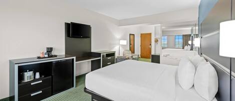A hotel room with a large bed, a flat-screen TV, a desk with a computer, a minibar, and a bathroom visible through an open door.