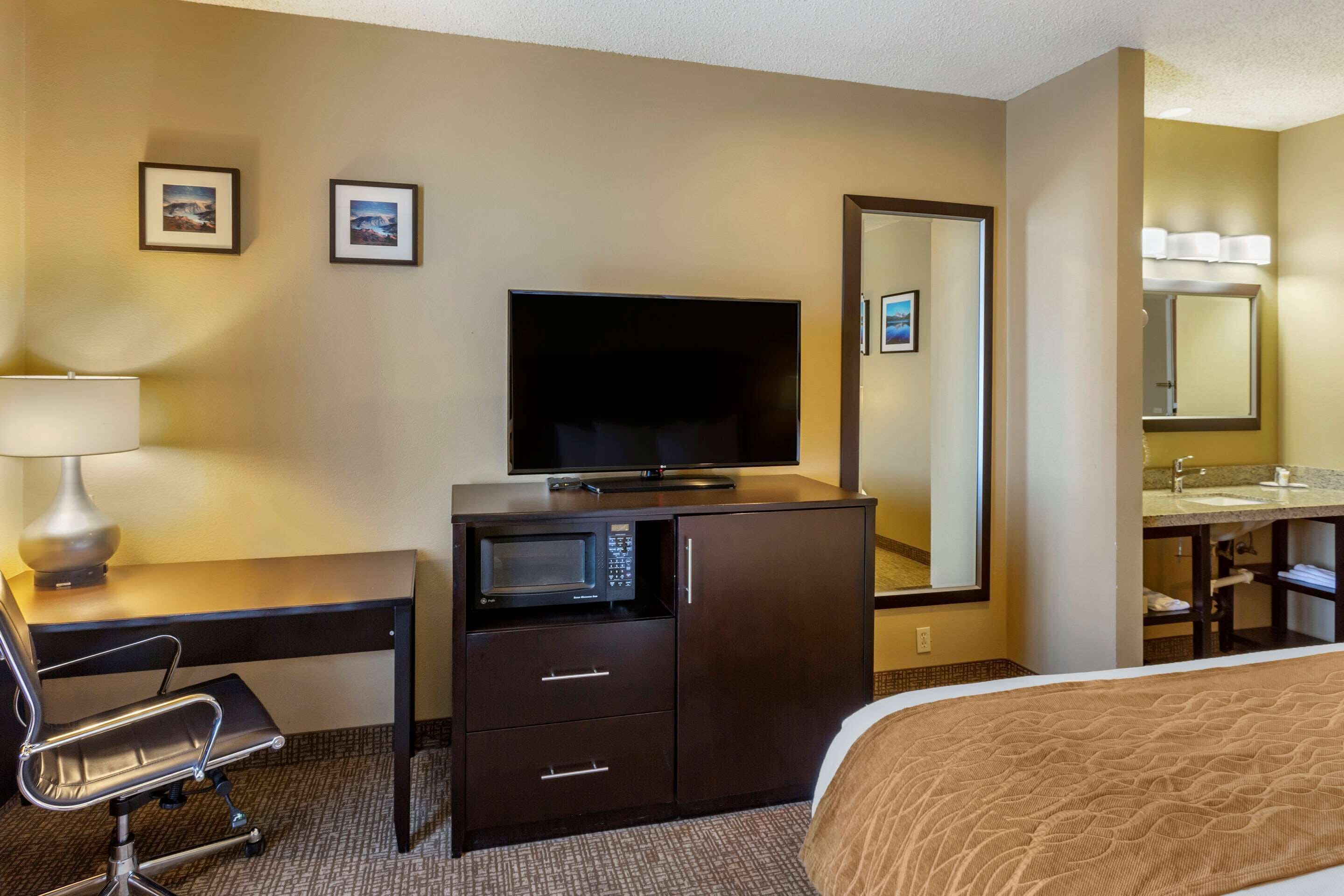A hotel room with a bed, a desk with a lamp, a television, a microwave, a mirror, and a bathroom with a sink and mirror.