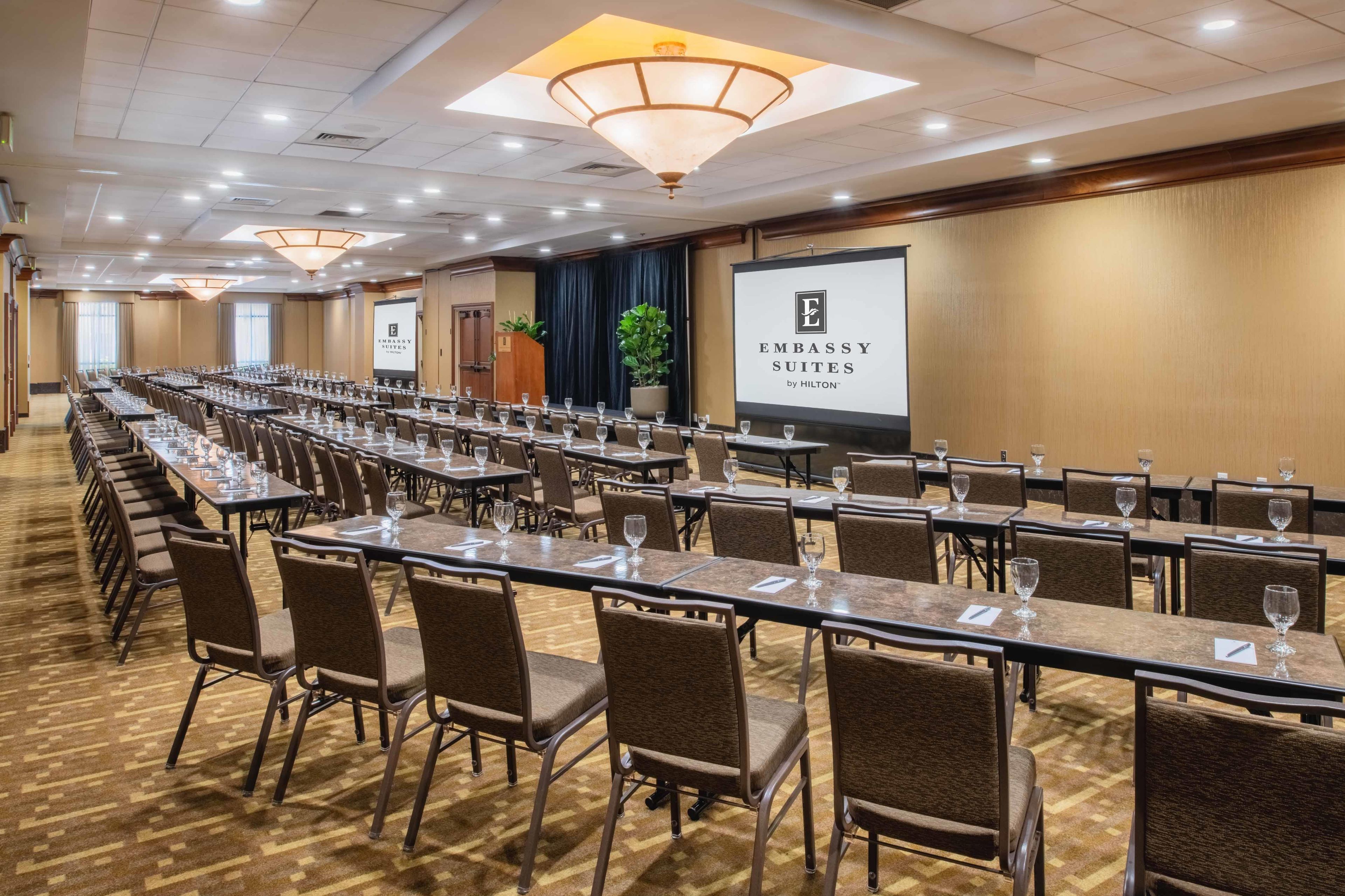 A conference room with long tables, chairs, and a projector screen displaying 'Embassy Suites by Hilton'.