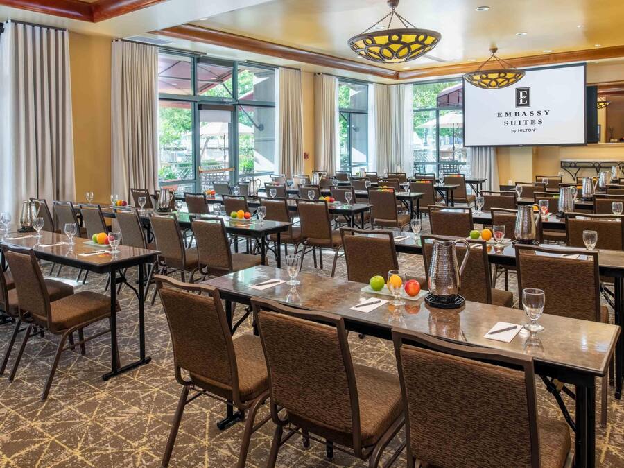 Uma sala de conferências com várias mesas, cadeiras e uma tela grande exibindo 'Embassy Suites by Hilton'.