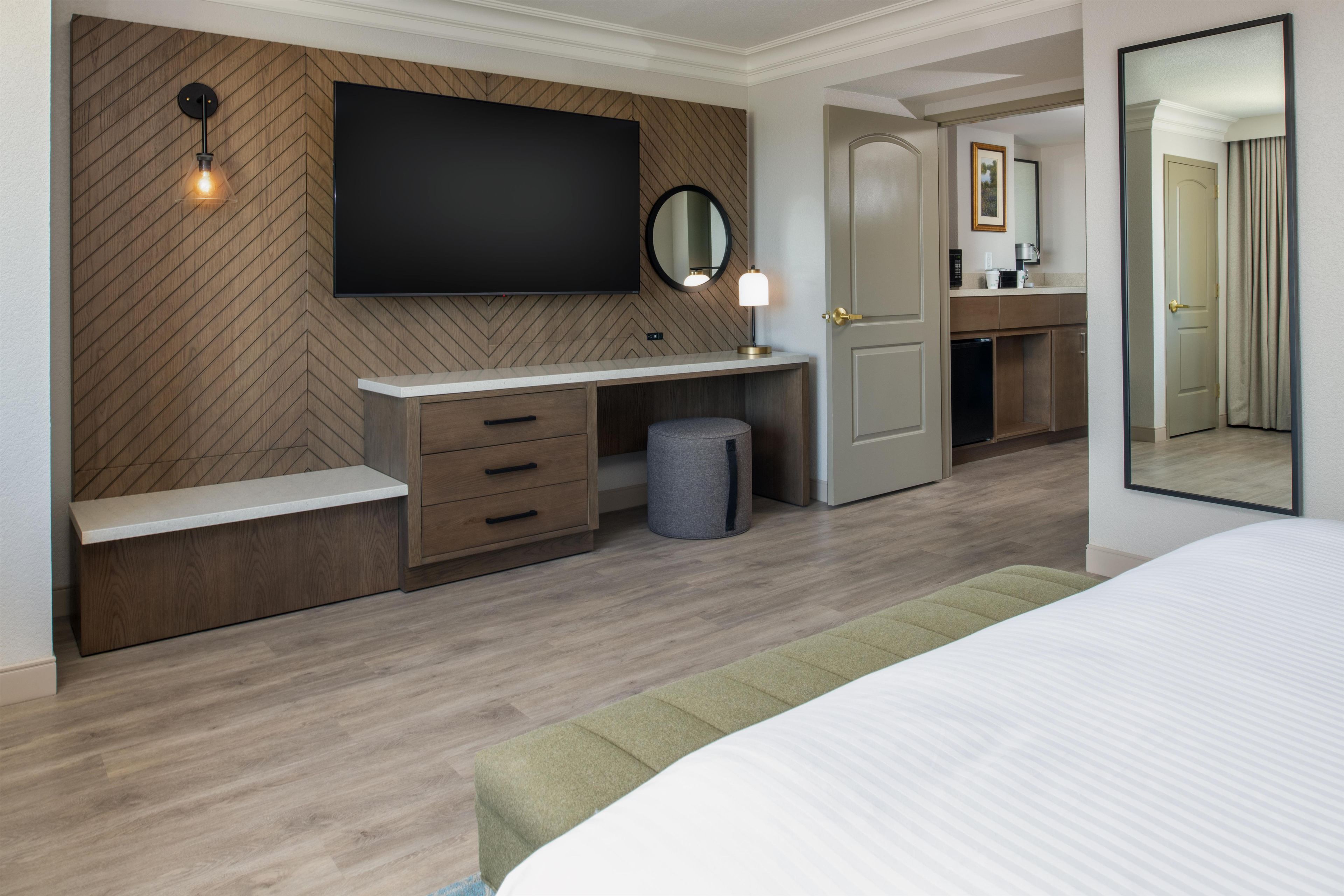 Una habitación de hotel moderna con un televisor de pantalla plana montado en una pared con estampado de espiga, una mesita de noche, un pequeño escritorio con lámpara, un cesto de basura y un espejo grande.