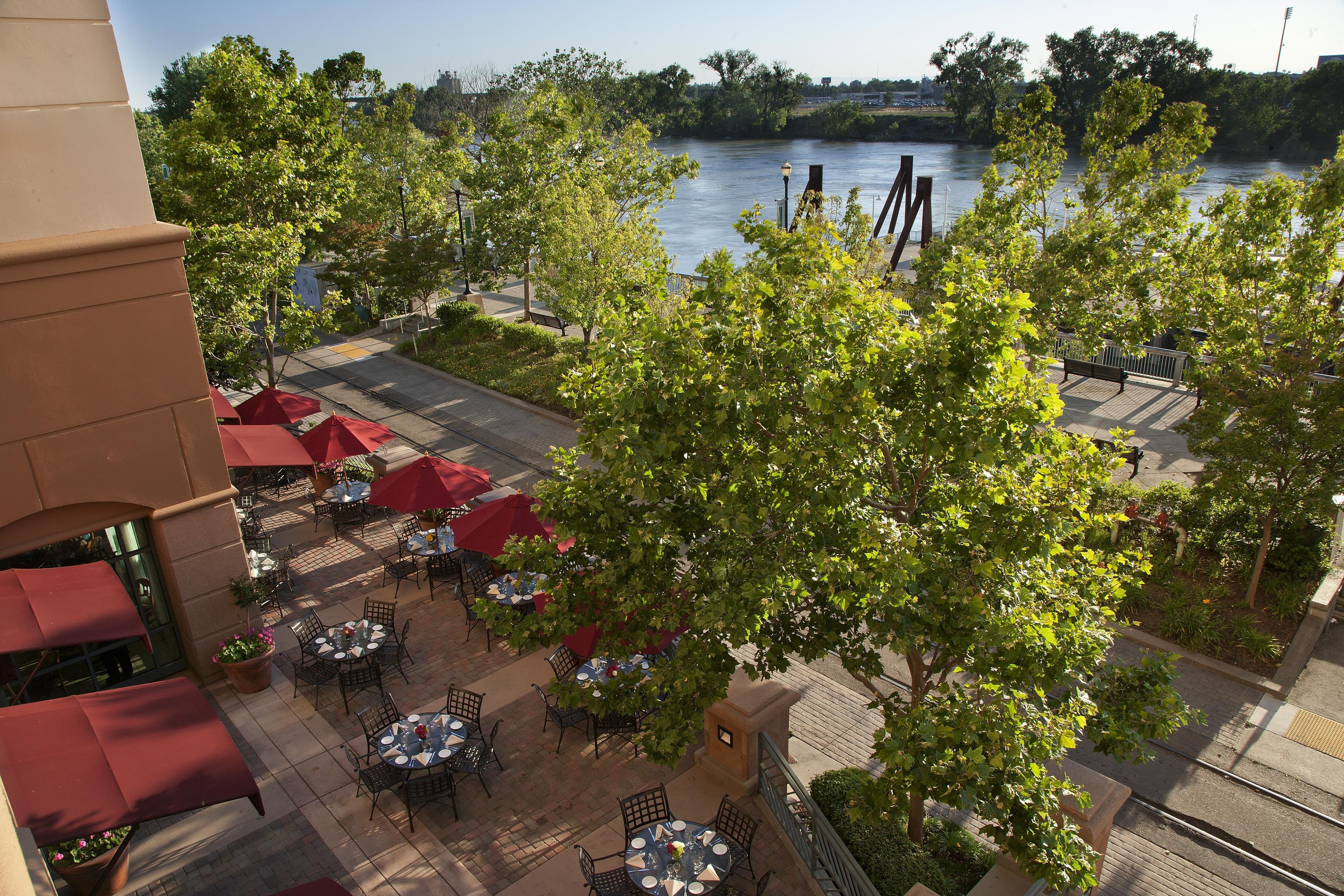 An outdoor dining area with tables and chairs, surrounded by trees and overlooking a river.