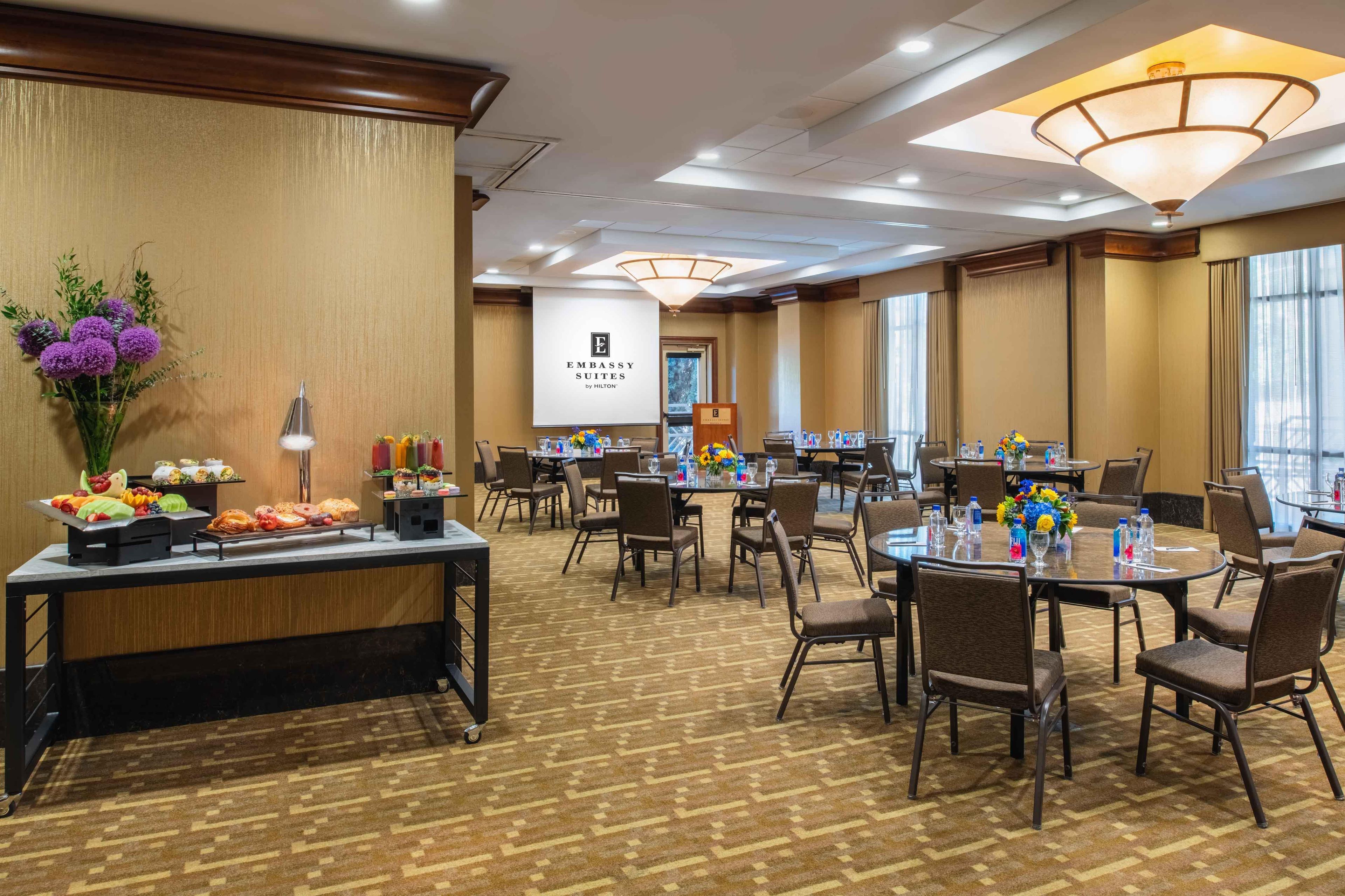 Un salón de conferencias de hotel con mesas redondas, sillas, una zona de buffet y una gran pantalla mostrando el nombre del hotel.