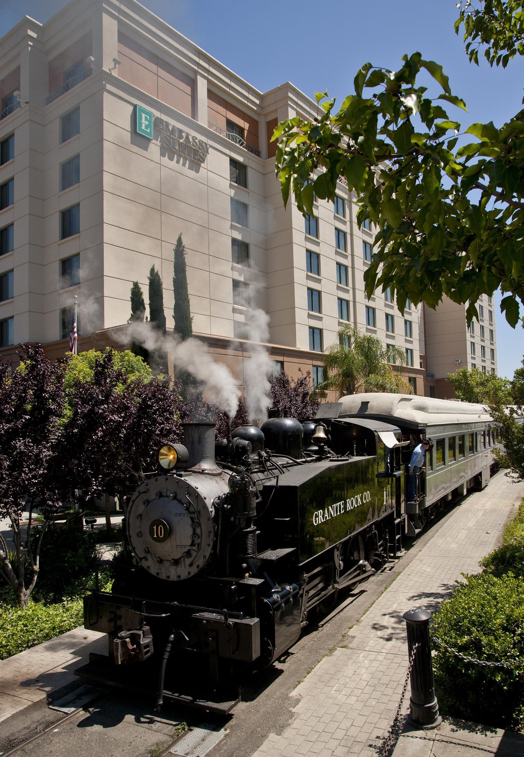 A steam locomotive with the number 10, labeled 'GRANITE ROCK CO.', on a track. In the background, there is a building with the sign 'EMBASSY SUITES'.