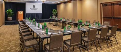 Un salón de conferencias con una larga mesa, sillas, un atril y una pantalla con el texto 'EMBASSY SUITES by HILTON'.