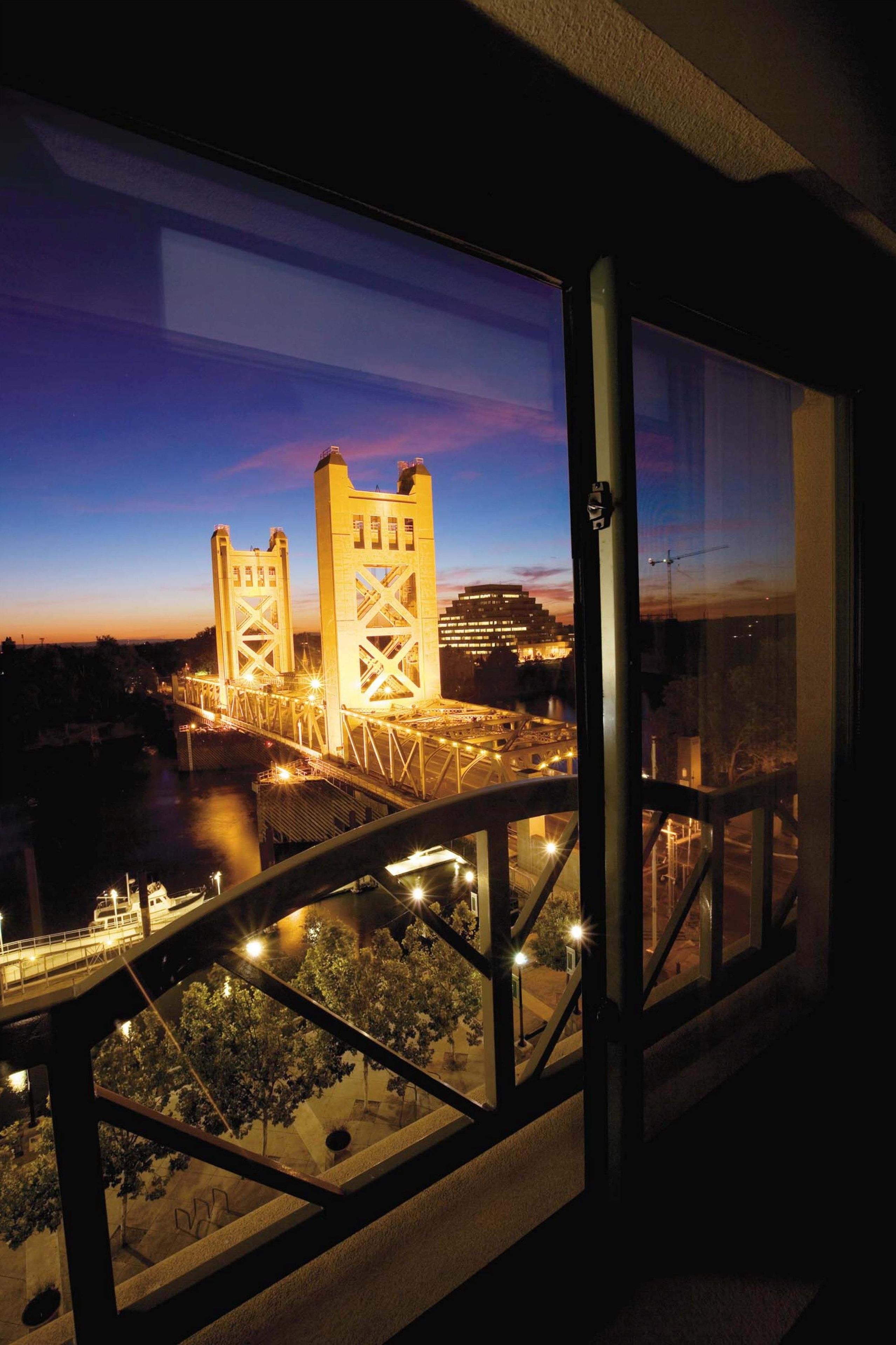 A nighttime view of a bridge with two towers, a river, and a cityscape in the background.