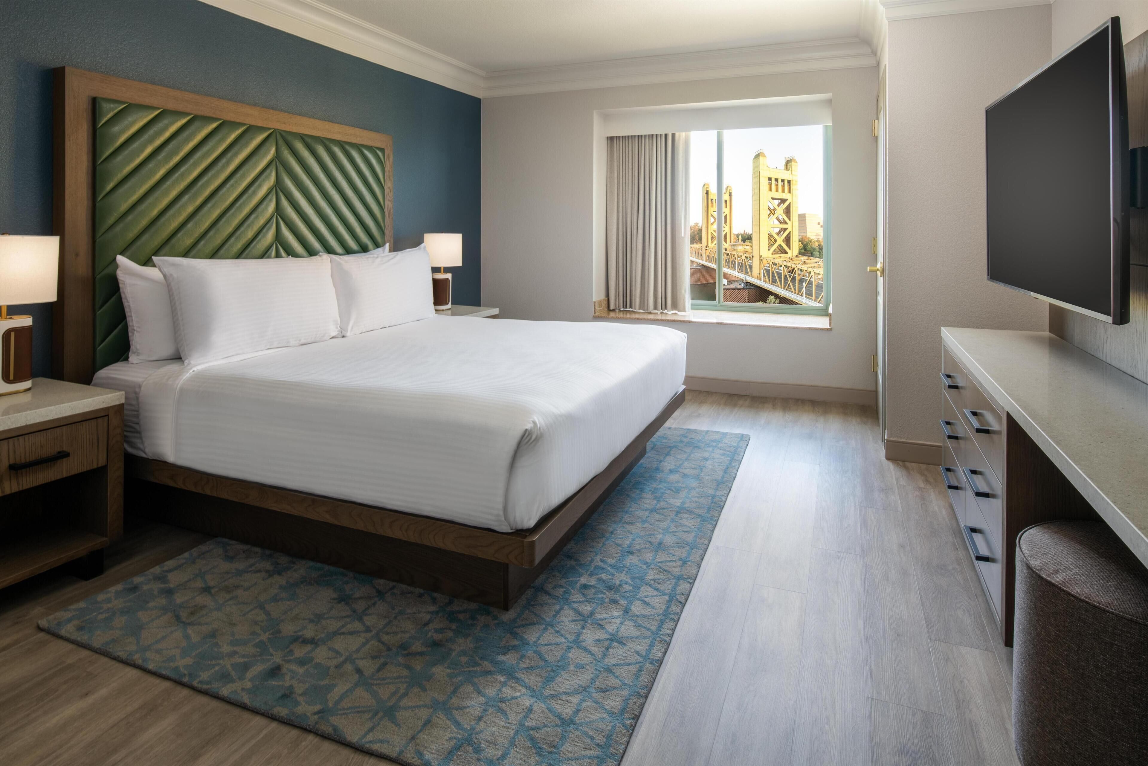 Een hotelkamer met een groot bed, een televisie, een raam met uitzicht op de stad en een bureau met lades.