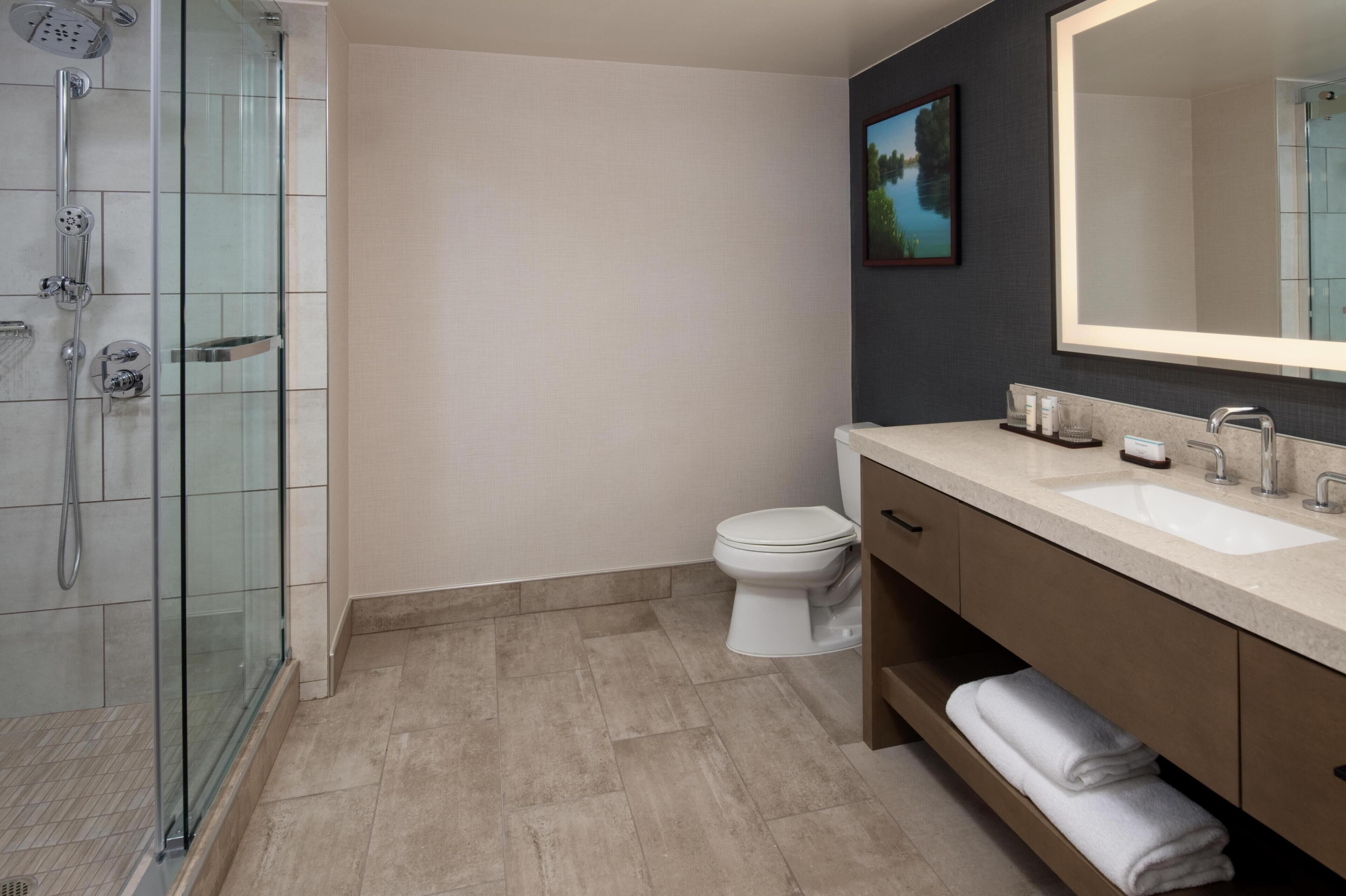 A modern bathroom with a glass-enclosed shower, a white toilet, and a large mirror above a beige vanity.