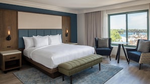 A hotel room with a large bed, a bench, two armchairs, a small table, and a view of a bridge and water.