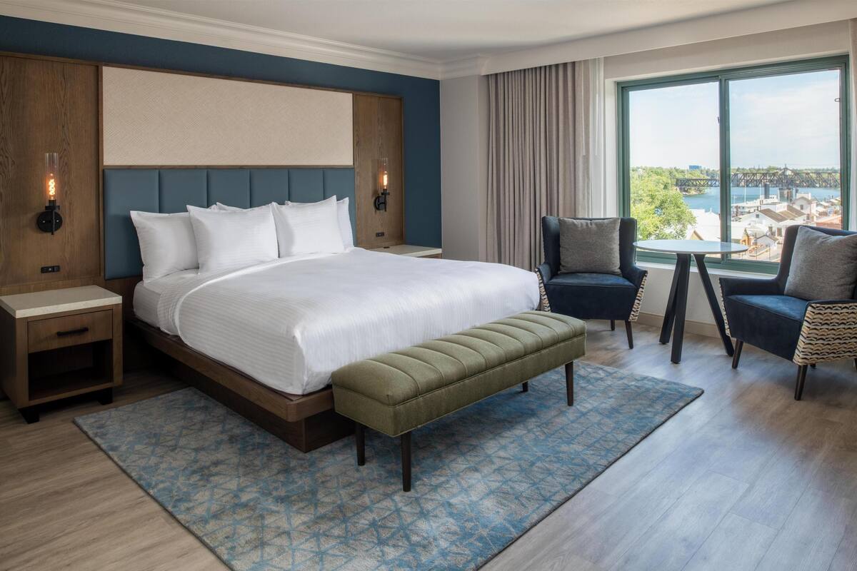 Habitación de hotel con una cama grande, un banco, dos sillones, una mesita y vistas a un puente y al agua.