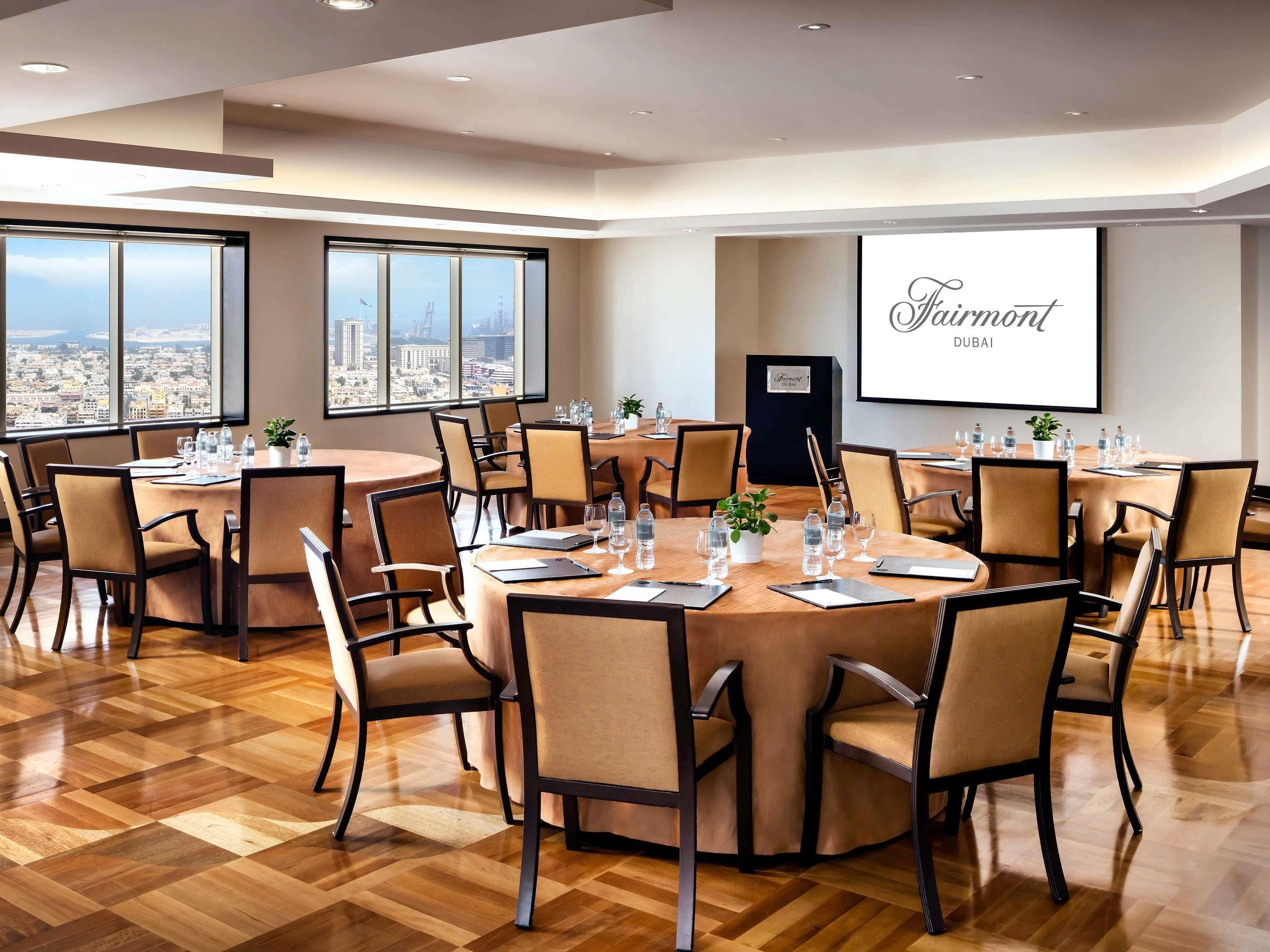 Un salón de conferencias con varias mesas, sillas y una pantalla grande que muestra 'Fairmont Dubai'.