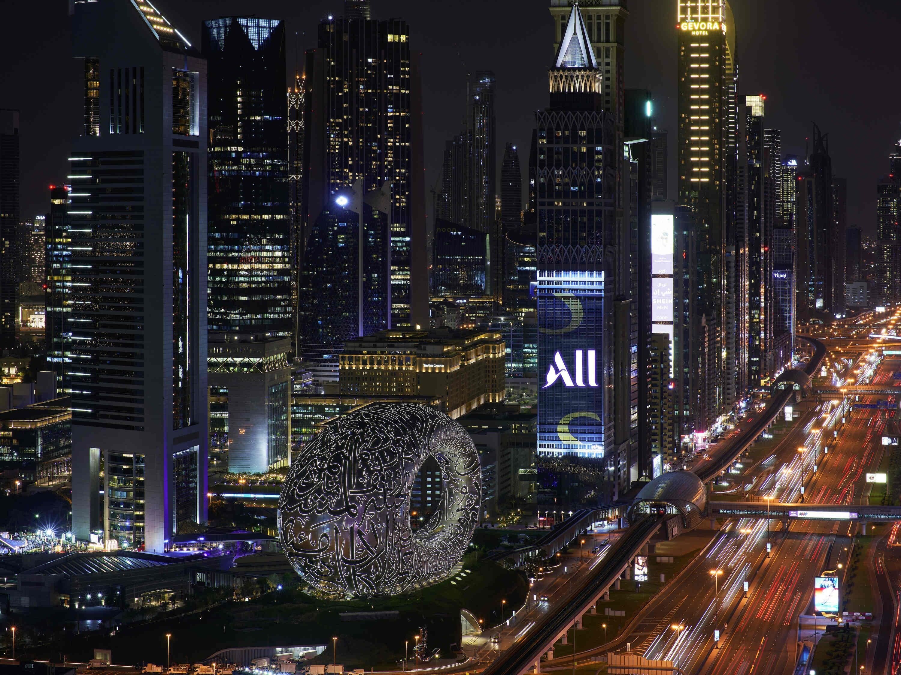 A cityscape with illuminated skyscrapers, including a prominent building with a unique spherical structure and a large billboard with the word 'All'.