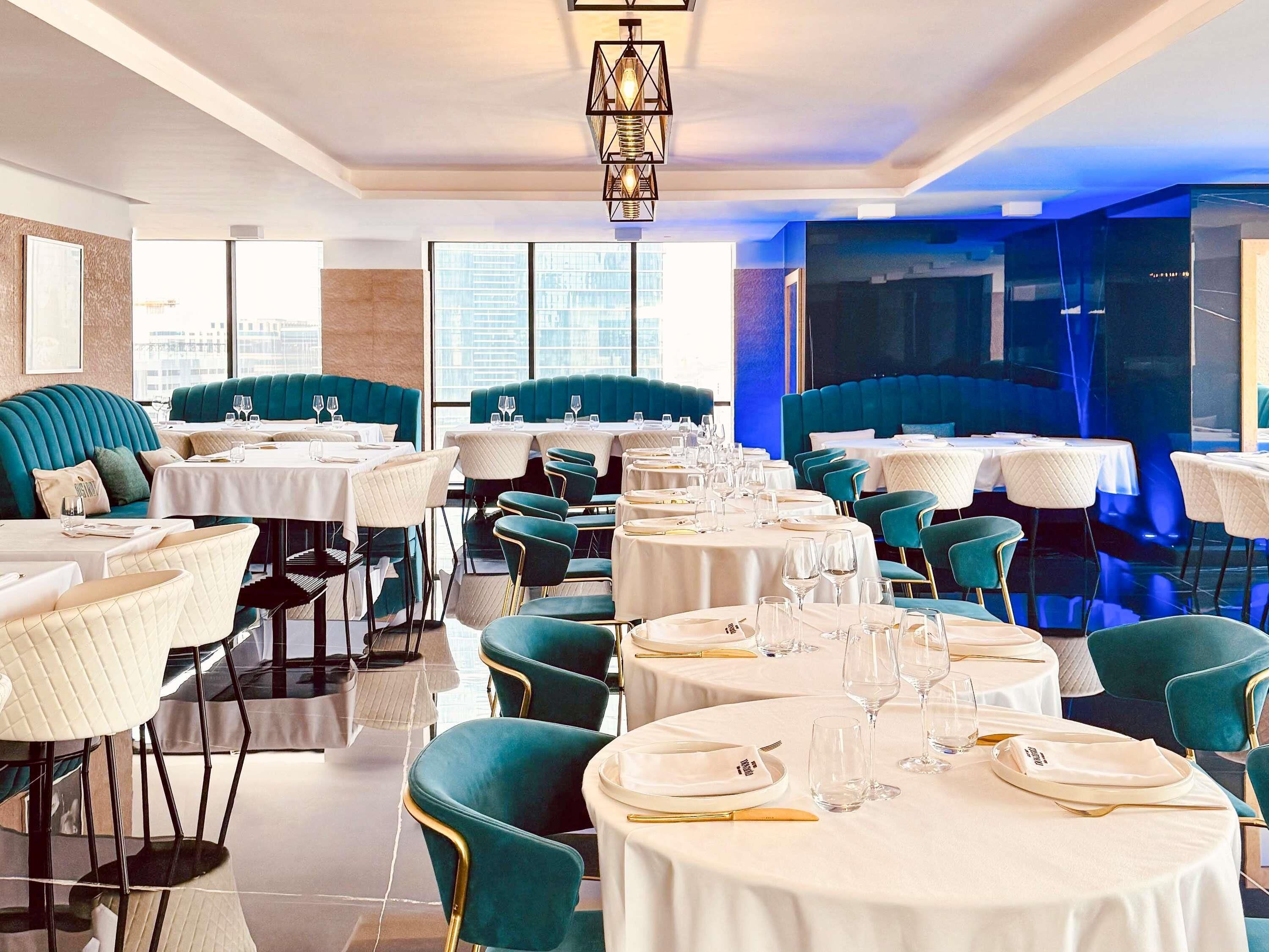 Un restaurant avec des tables rondes, des banquettes recouvertes d’un tissu turquoise et un lustre moderne.