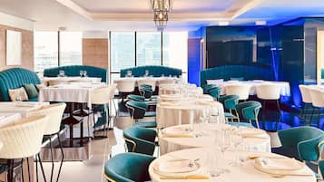 Un restaurant avec des tables rondes, des banquettes recouvertes d’un tissu turquoise et un lustre moderne.