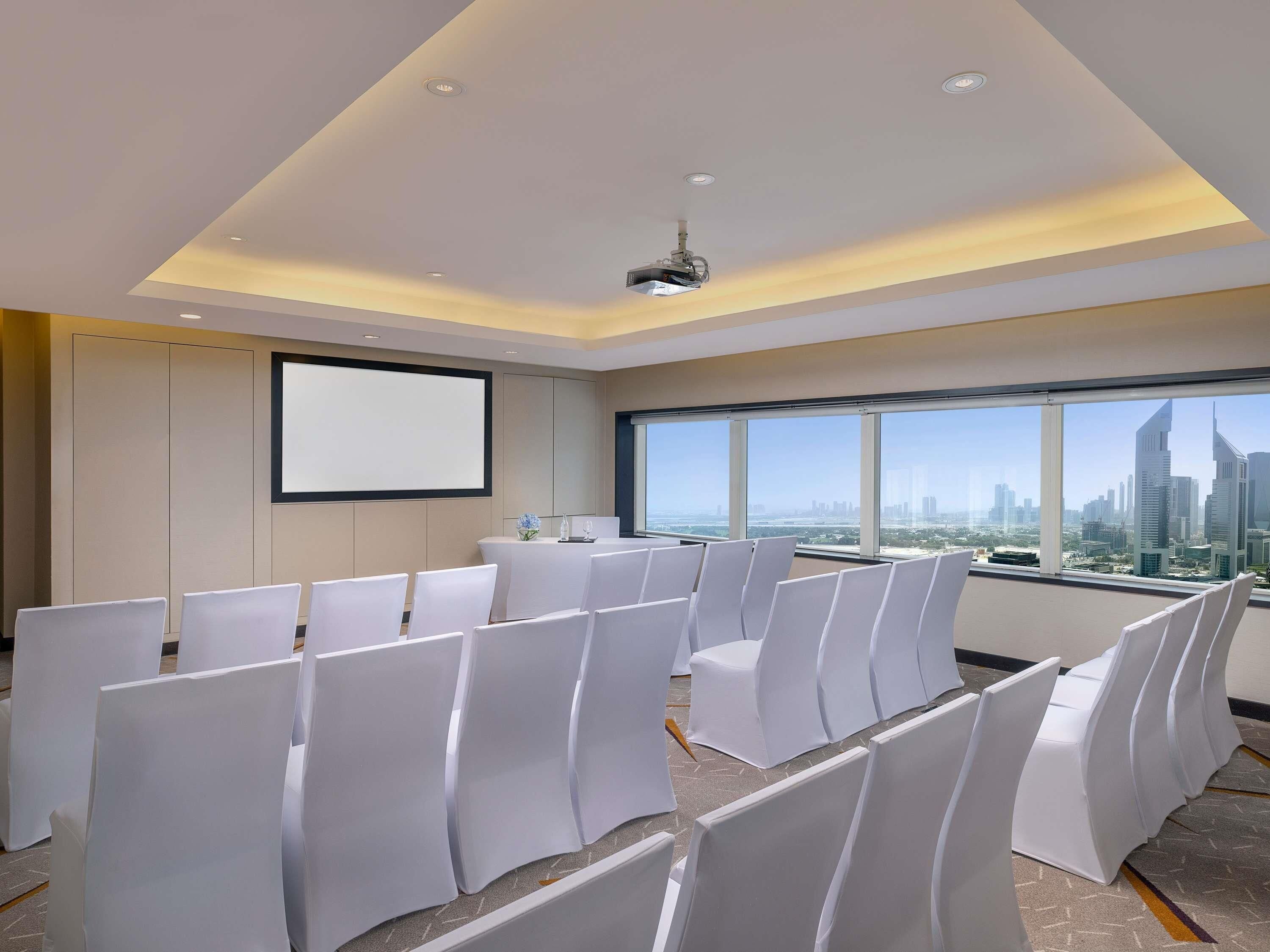 Une salle de conférence dotée d’un grand écran, de rangées de chaises blanches et offrant une vue sur la ville grâce à de grandes fenêtres.