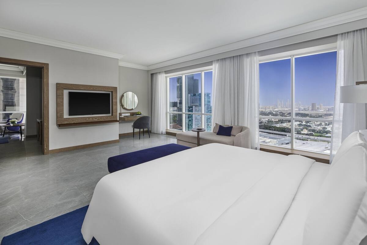 Ein Hotelzimmer mit einem großen Bett, einem Schreibtisch, einem Stuhl, einem Fernseher und Blick auf die Stadt.