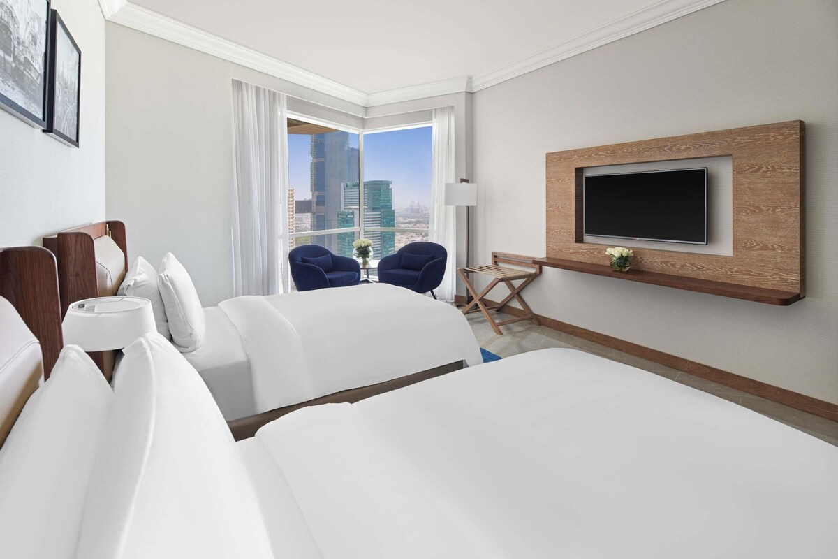 A hotel room with a large bed, two armchairs, a TV mounted on a wooden panel, and a city view.