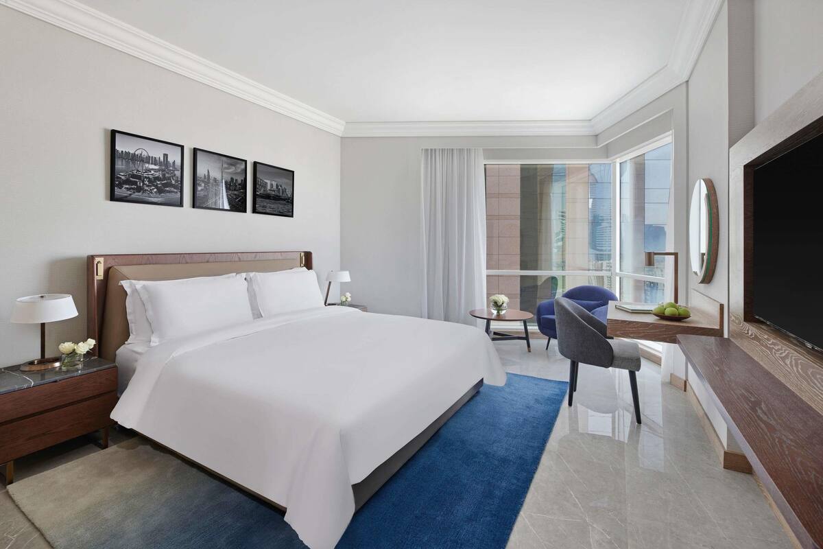 Ein Hotelzimmer mit einem großen Bett, einem Schreibtisch, einem Sessel, einem Fernseher und einem Fenster mit Blick auf die Stadt.