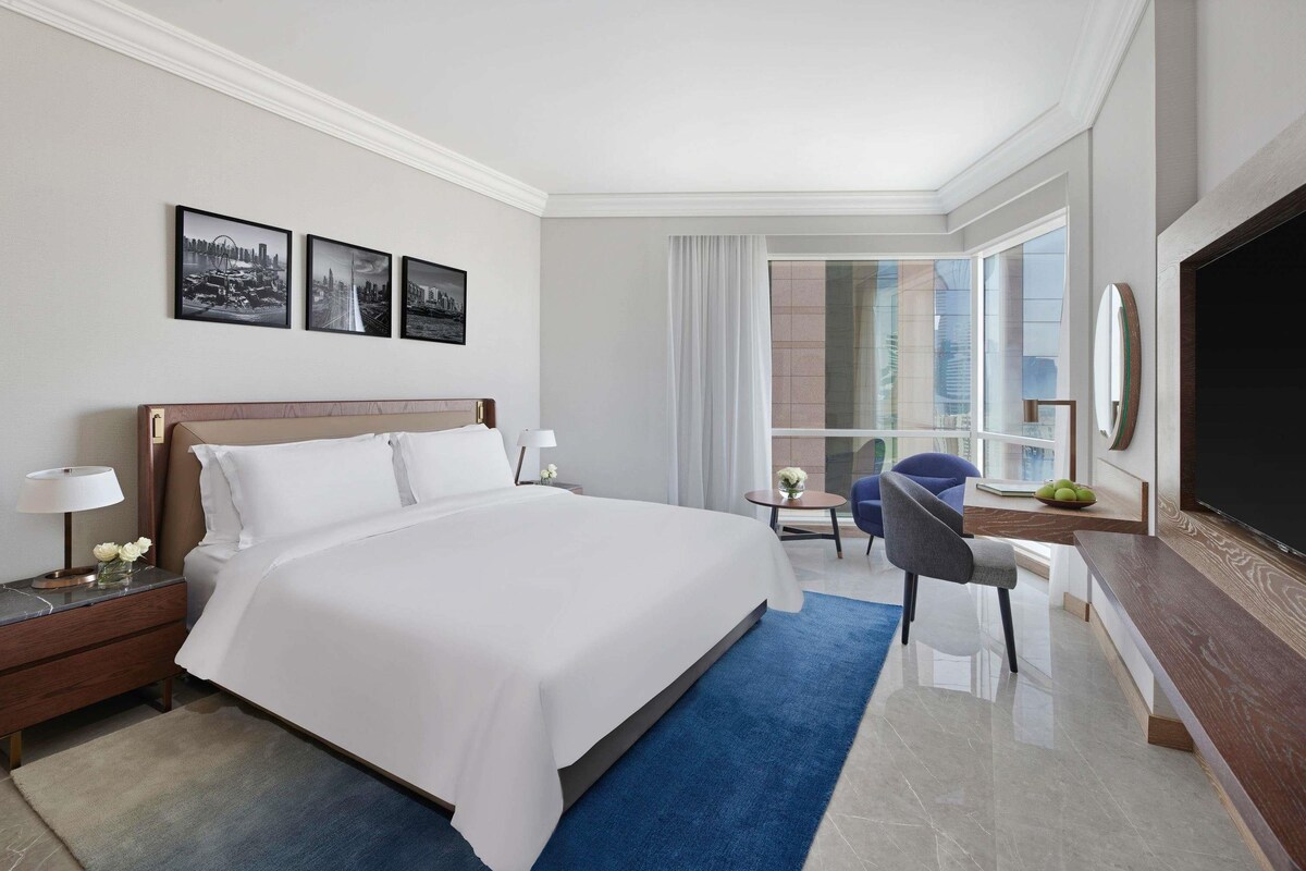 A hotel room with a large bed, a desk, a chair, a TV, and a window with a city view.