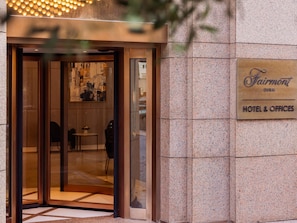 A hotel entrance with a glass door, a sign reading 'Fairmont Dubai Hotel & Offices,' and a view into the lobby area.